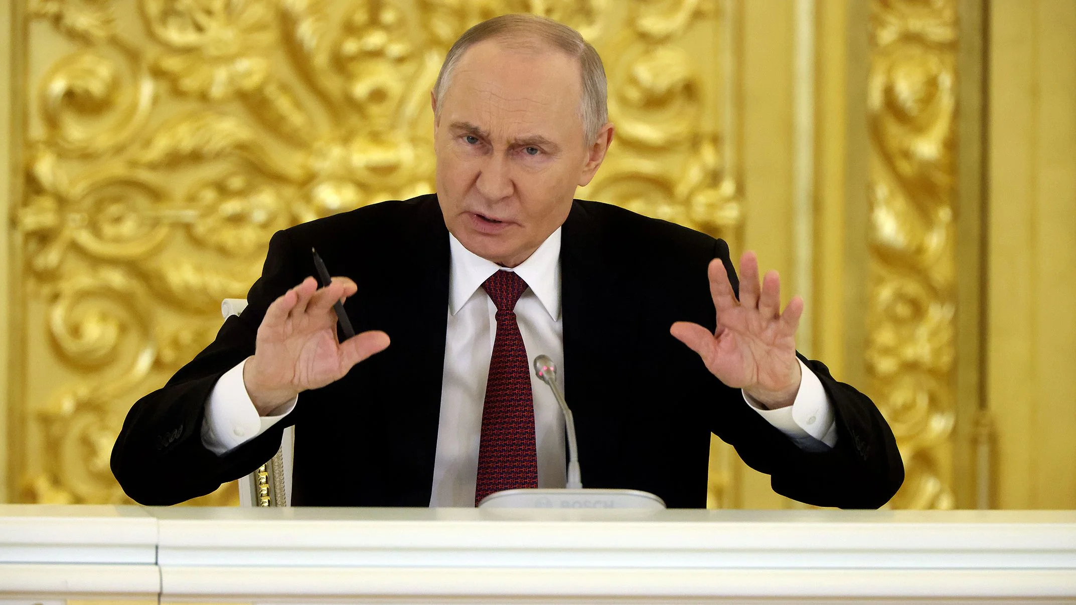 Володимир Путін жестикулює обома руками під час виступу за столом у залі з золотим оздобленням